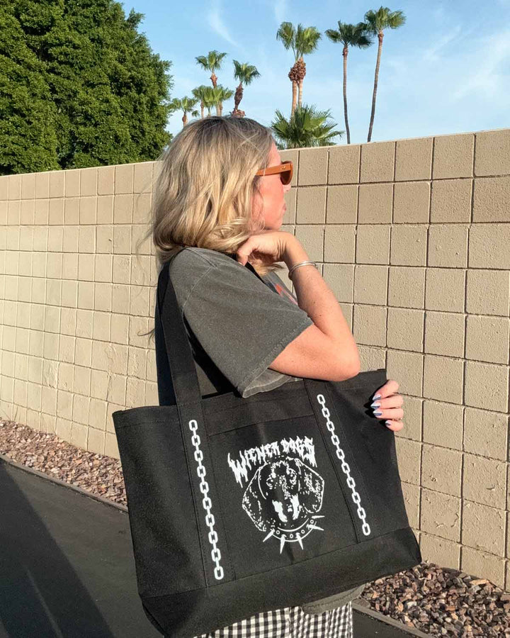 A woman wearing sunglasses and a gray t-shirt carries a large black canvas tote bag with a white graphic print featuring the text 'WIENER DOGS' and a circular design. She is standing outdoors against a beige brick wall with palm trees in the background.