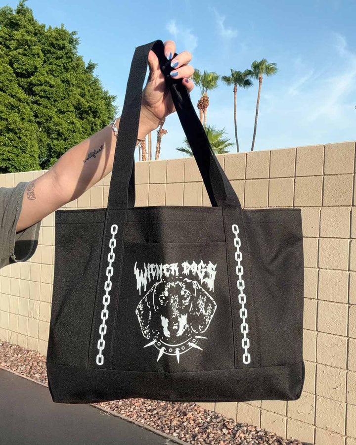 A black canvas tote bag with white graphic print of a dog head and the text 'WIENER DOGS' held by a person's hand outdoors in front of a beige brick wall and palm trees.