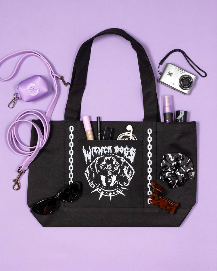 A black tote bag with a white 'WIENER DOGS' graphic, styled in a flat lay on a lavender background with various accessories including a purple phone case, sunglasses, a camera, and a purple strap.