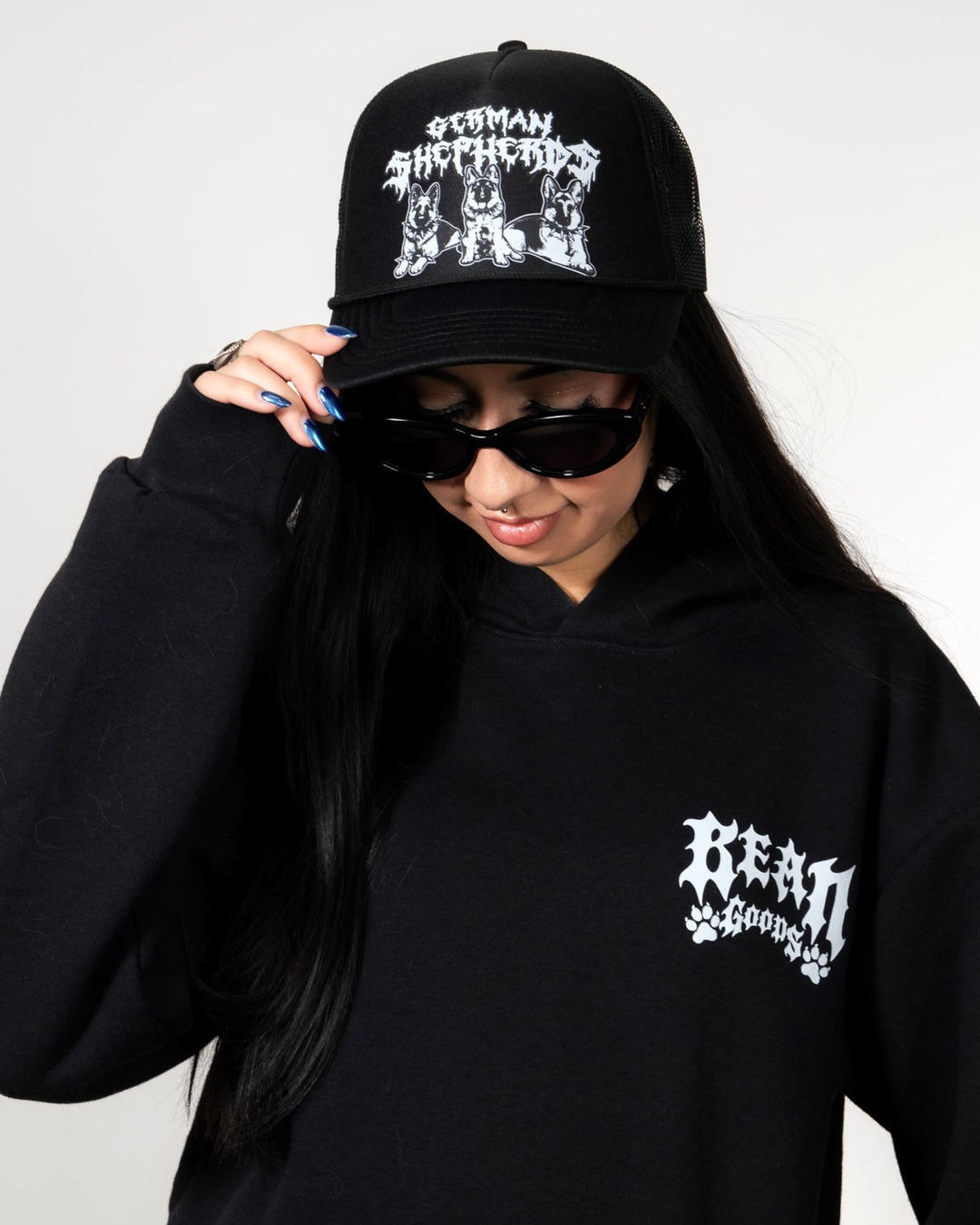 A person wearing a black baseball cap with a white gothic-style graphic and text, a black hoodie with a matching white chest graphic, and black sunglasses, posed against a light gray background.