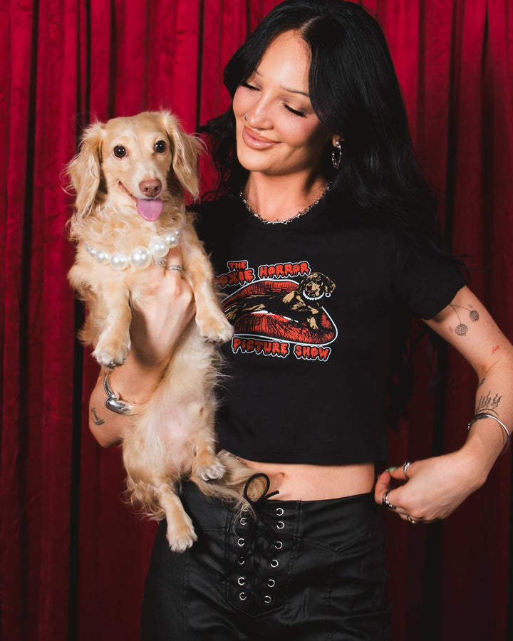 A woman posing in front of a red velvet curtain, wearing a black short-sleeve crop top with a graphic print that reads 'The Doxie Horror Picture Show' and black lace-up pants.