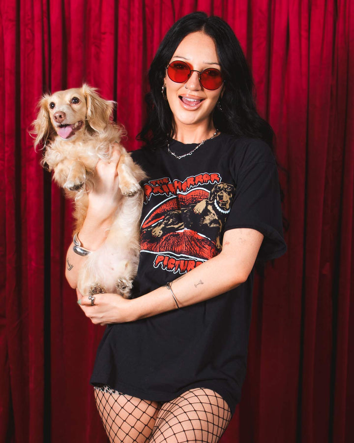 A woman modeling an oversized black graphic T-shirt with a red and beige 'The Doxie Horror Picture Show' lips design, posed in front of a red curtain backdrop.