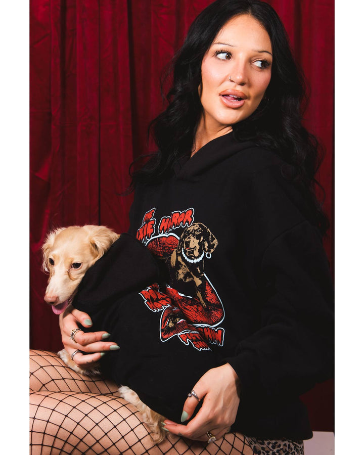 A woman stands in front of a red curtain wearing a black hoodie with a large graphic print on the front that reads 'The Doxie Horror Picture Show' with a stylized horror character. She is also wearing leopard-print shorts and black fishnet stockings.