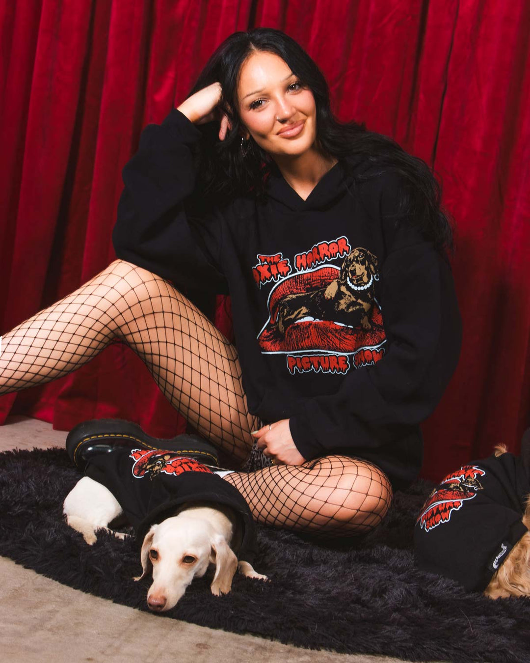 A woman stands in front of a red curtain wearing a black hoodie with a large graphic print on the front that reads 'The Doxie Horror Picture Show' with a stylized horror character. She is also wearing leopard-print shorts and black fishnet stockings.