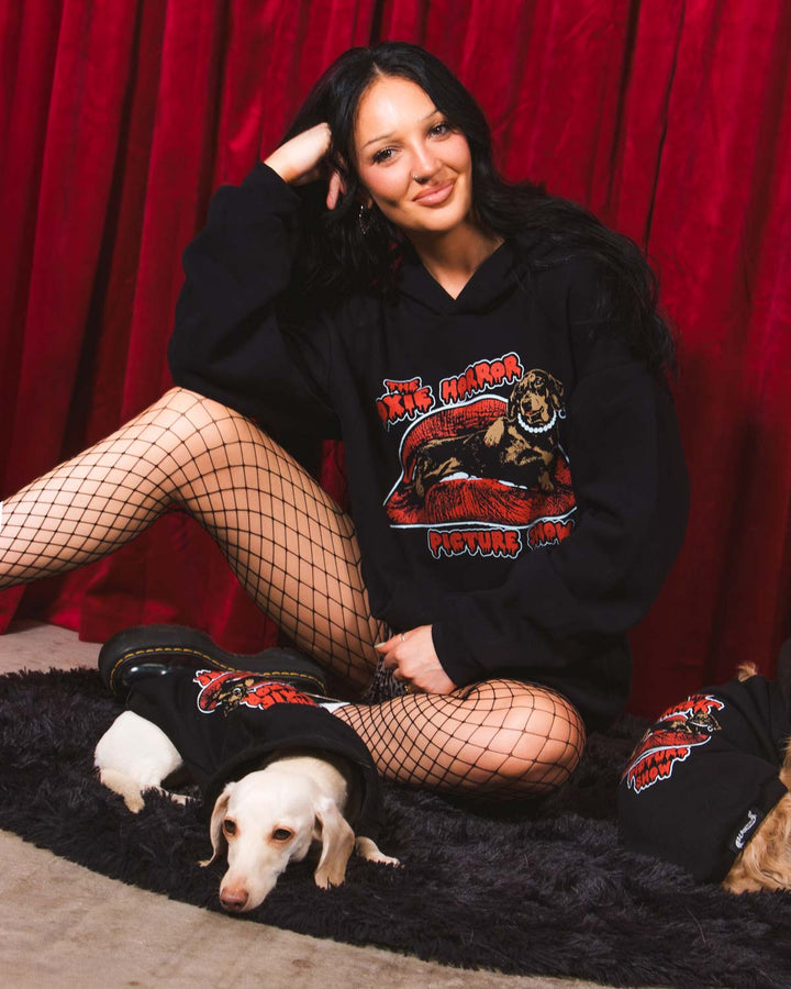 A woman stands in front of a red curtain wearing a black hoodie with a large graphic print on the front that reads 'The Doxie Horror Picture Show' with a stylized horror character. She is also wearing leopard-print shorts and black fishnet stockings.