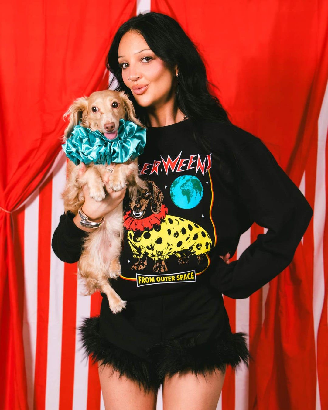 A woman holding a small tan dog in front of a red and white striped curtain backdrop. The woman is wearing a black long-sleeve graphic sweatshirt with a colorful space-themed print and black feather-trimmed shorts.