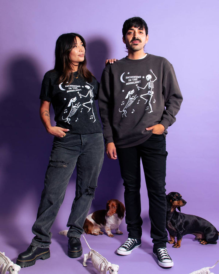 A man and a woman pose together wearing matching black graphic sweatshirts featuring white skeleton illustrations and the text 'DACHSHUND IN THE MOONLIGHT'. They are standing against a solid purple studio background with two dachshunds at their feet.