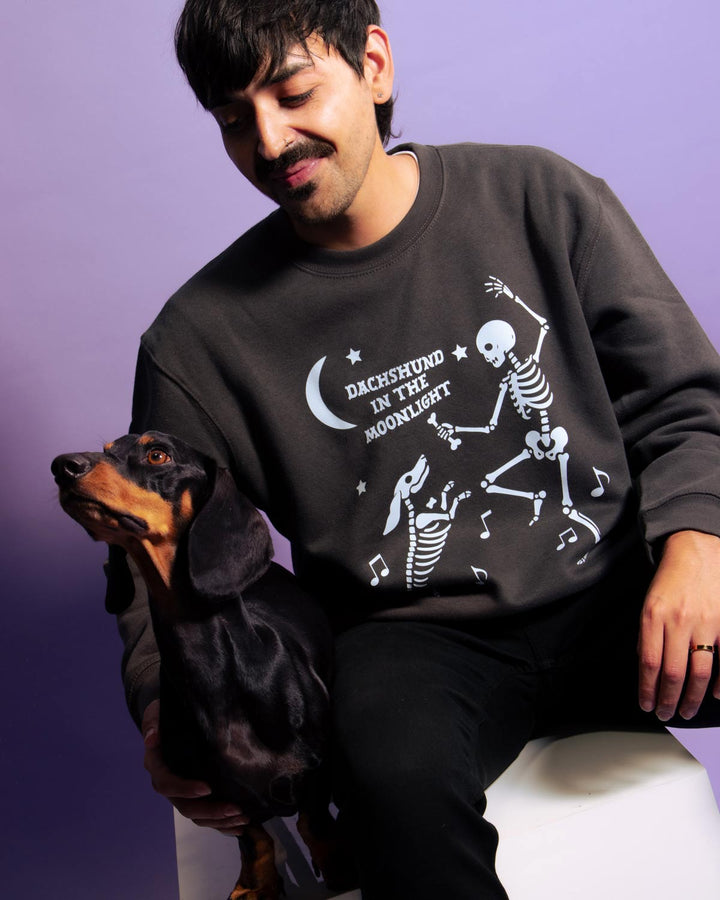 A man wearing a dark charcoal gray sweatshirt with a white graphic of dancing skeletons and the text 'DACHSHUND IN THE MOONLIGHT' sits on a white stool next to a black and tan dachshund against a purple background.