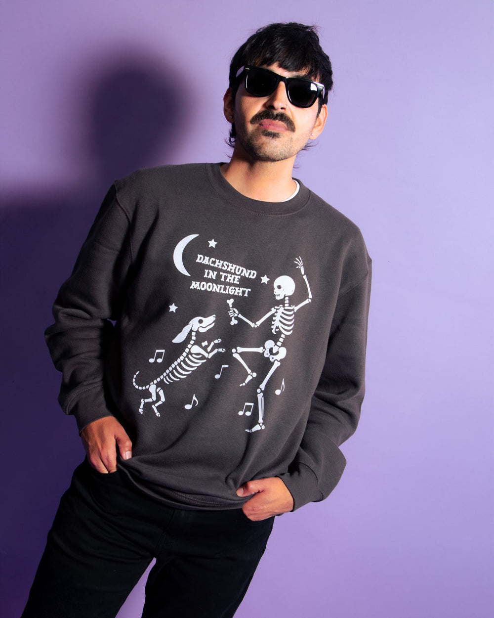 A man wearing a dark gray crewneck sweatshirt with a white graphic of dancing skeletons and the text 'DACHSHUND IN THE MOONLIGHT' against a solid purple background.