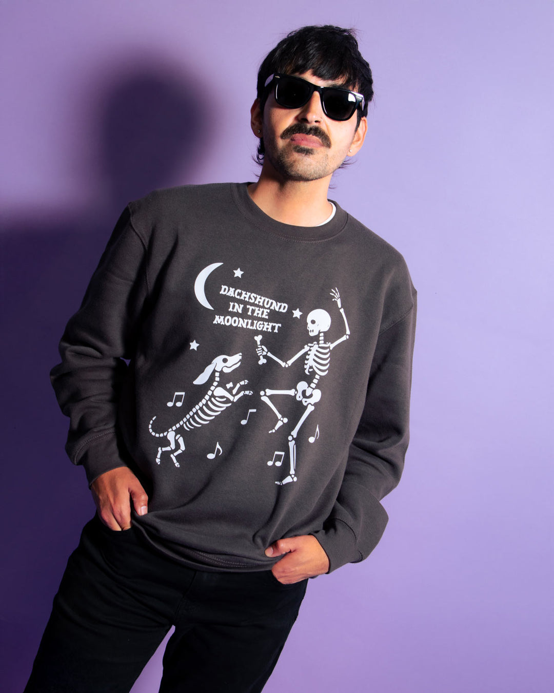 A man wearing a dark gray crewneck sweatshirt with a white graphic of dancing skeletons and the text 'DACHSHUND IN THE MOONLIGHT' against a solid purple background.