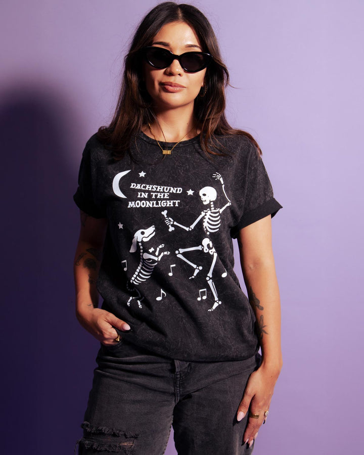 A woman modeling a dark charcoal gray graphic t-shirt with a white print of dancing skeletons and the text 'DACHSHUND IN THE MOONLIGHT' against a solid purple background.