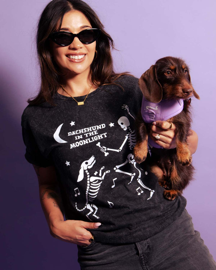 A woman wearing a black graphic t-shirt with a white 'Dachshund in the Moonlight' skeleton dog design, holding a small brown dachshund wearing a purple harness against a solid purple background.