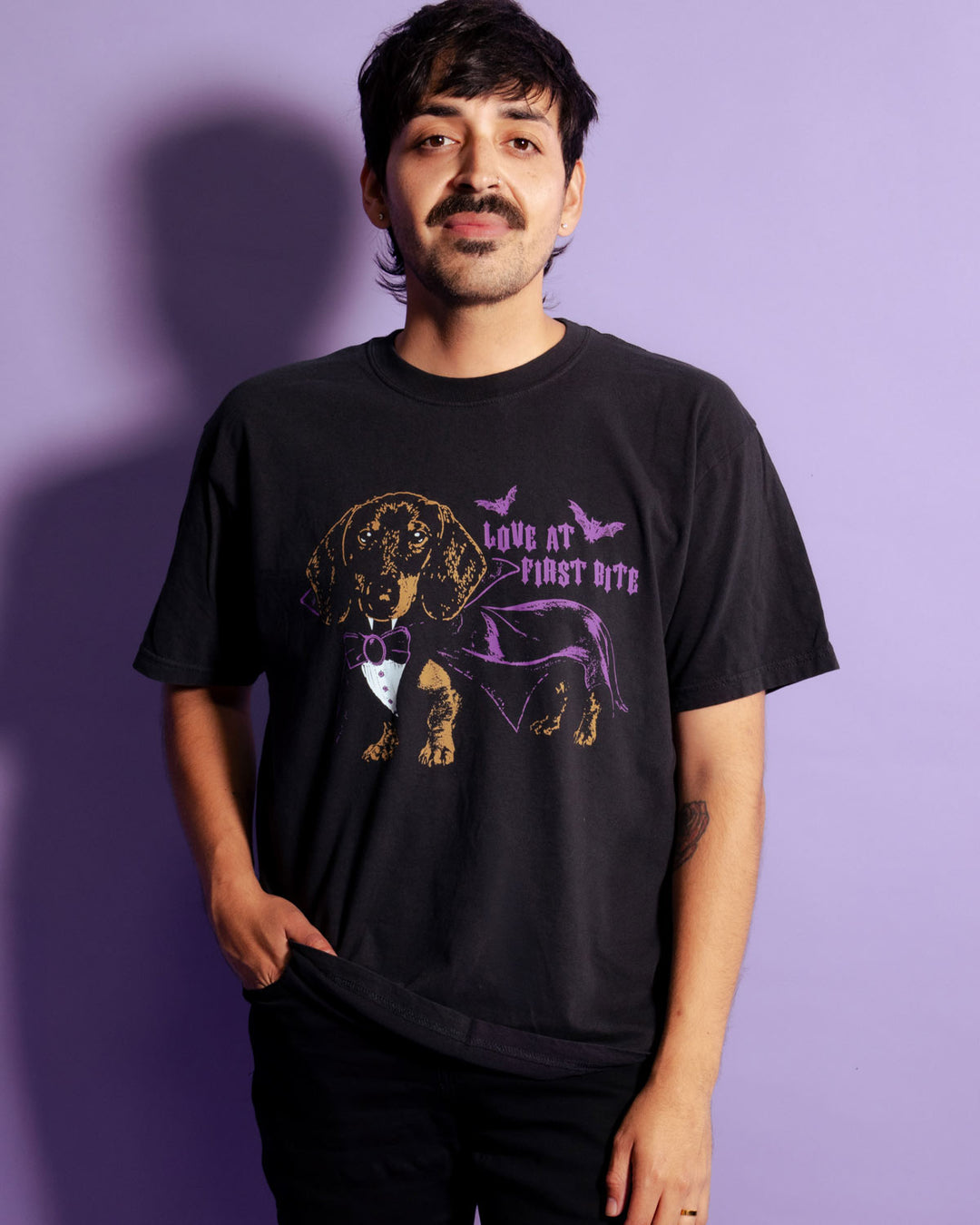 A man wearing a black graphic t-shirt with a purple and gold dachshund design and the text 'LOVE AT FIRST BITE' against a solid purple background.