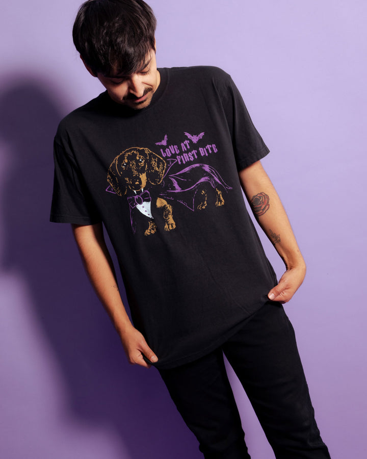 A man wearing a black graphic t-shirt with a purple and gold dachshund design and the text 'LOVE AT FIRST BITE', posed against a solid purple background.