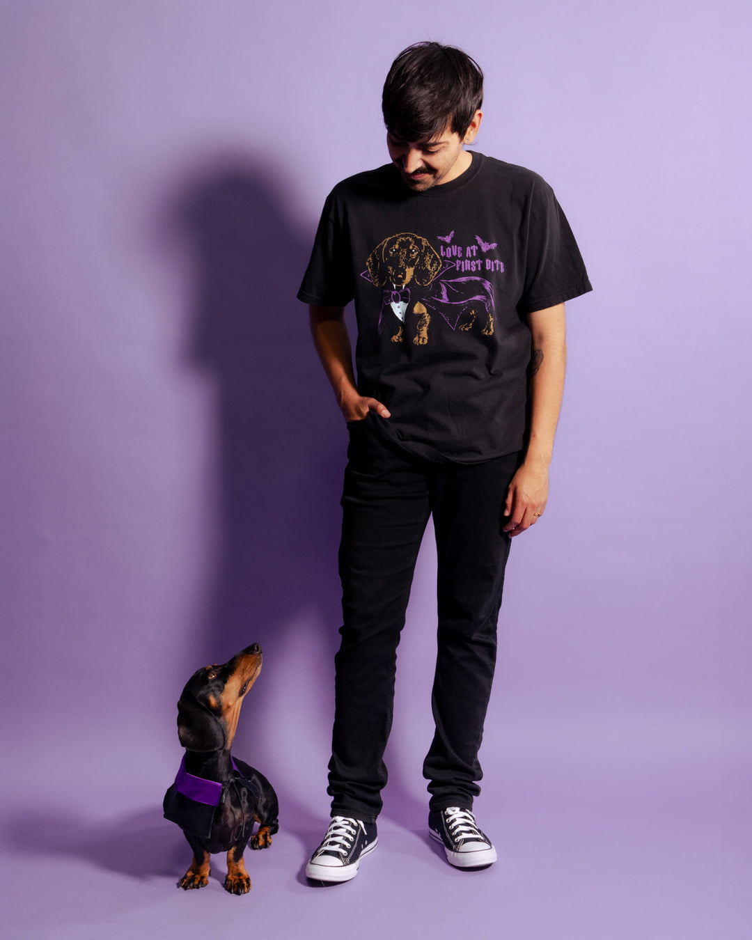 A man wearing a black graphic t-shirt with a dog design and the text 'LOVE AT FIRST BITE', paired with black pants and sneakers, standing next to a dachshund wearing a purple harness against a solid purple background.