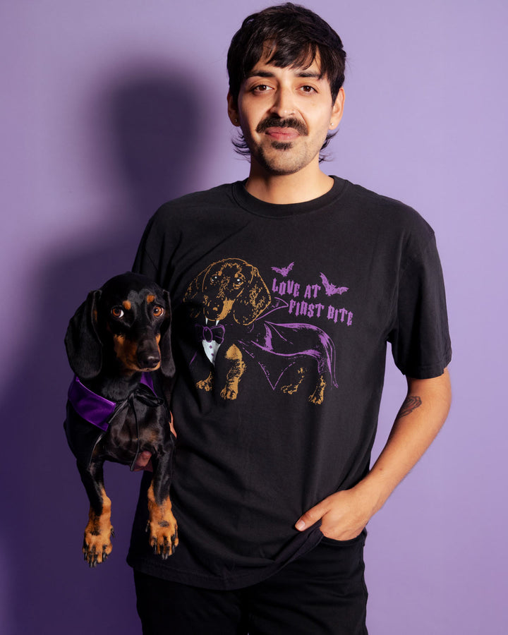 A man wearing a black graphic t-shirt with a dachshund wearing a purple cape, standing against a solid purple background. The t-shirt features a graphic of a dachshund wearing a purple cape and bowtie with the text 'Love at first bite'.