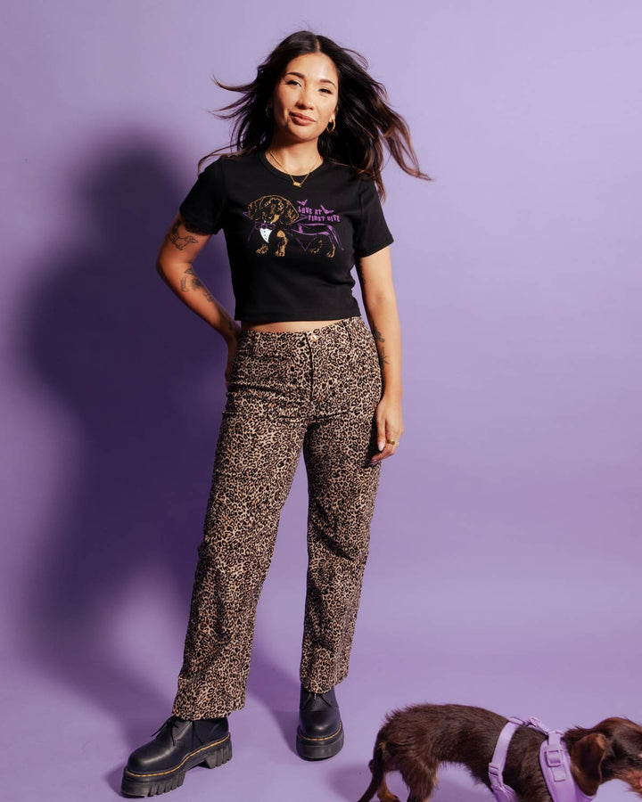 A woman posing in a black graphic t-shirt and leopard-print pants against a solid purple background, with a small brown dog wearing a purple harness in the lower right corner.