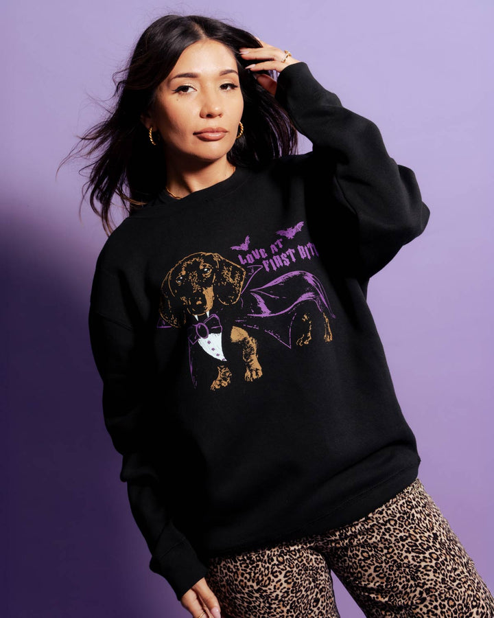 A woman modeling a black crewneck sweatshirt with a graphic of a dachshund wearing a purple cape and bow tie, with the text 'LOVE IT FIRST BITE'. She is wearing leopard print pants against a solid purple studio background.