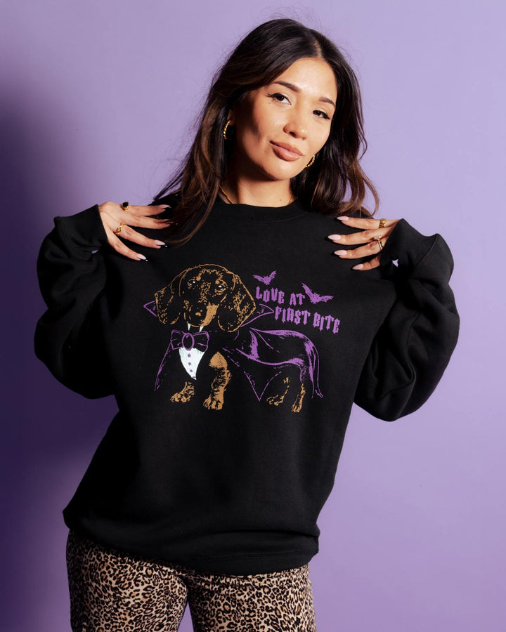 A woman modeling a black crewneck sweatshirt with a graphic of a dachshund dressed as a vampire wearing a purple cape and bowtie, with the text 'LOVE AT FIRST BITE'. She is wearing leopard print pants against a solid purple studio background.