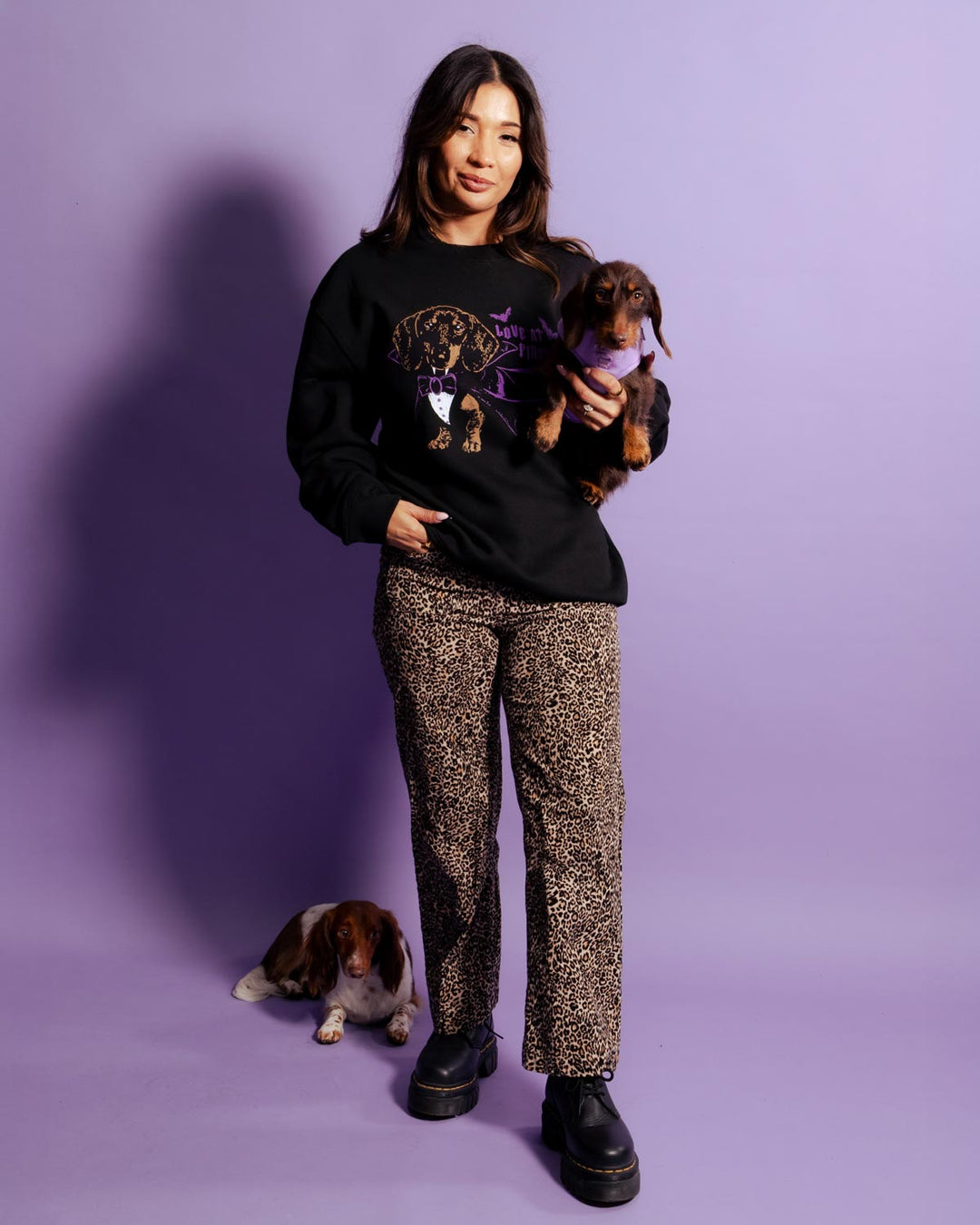 A woman stands against a lavender studio backdrop wearing a black graphic sweatshirt, leopard-print pants, and black boots, while holding a small brown dachshund. A second dog sits on the floor nearby.