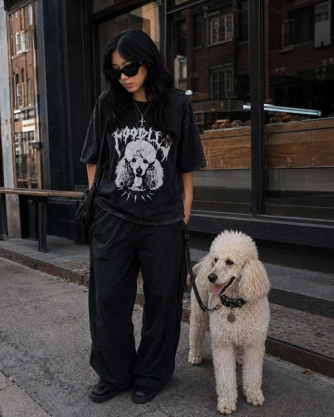 A woman wearing a black graphic t-shirt with a white dog print, black wide-leg pants, and black shoes, standing on a city sidewalk next to a fluffy white poodle. The background shows a storefront window.