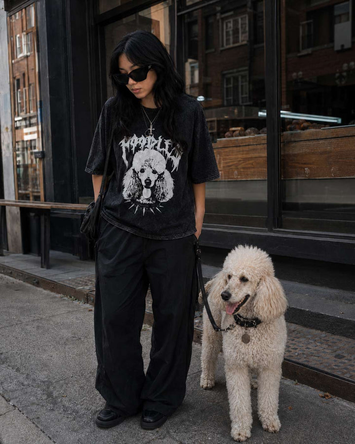 A woman wearing a black graphic t-shirt with a white dog print, black wide-leg pants, and black shoes, standing on a city sidewalk next to a fluffy white poodle. The background shows a storefront window.