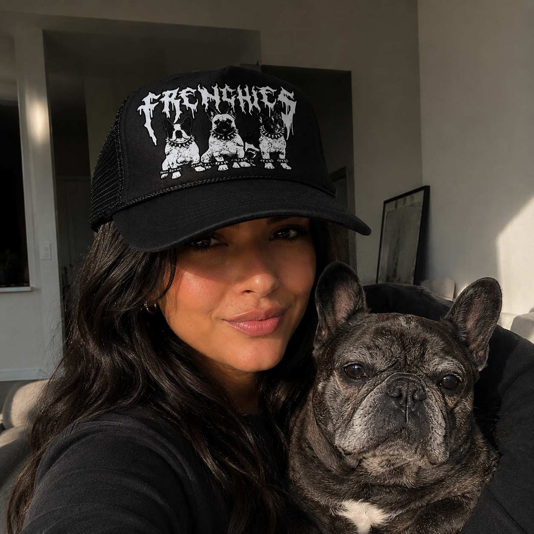 Woman wearing a black cap with 'FRENCHIES' and a dog's illustration, holding a French Bulldog.