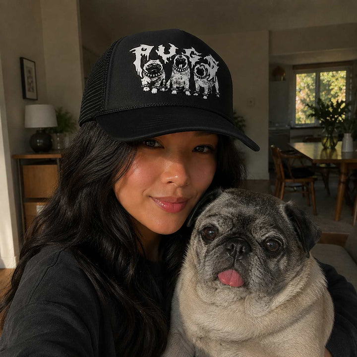 Woman wearing a black cap with a dog graphic, holding a pug in a home setting.